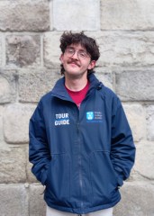 A male tour guide wearing a navy jacket with a light grey brick wall behind him.