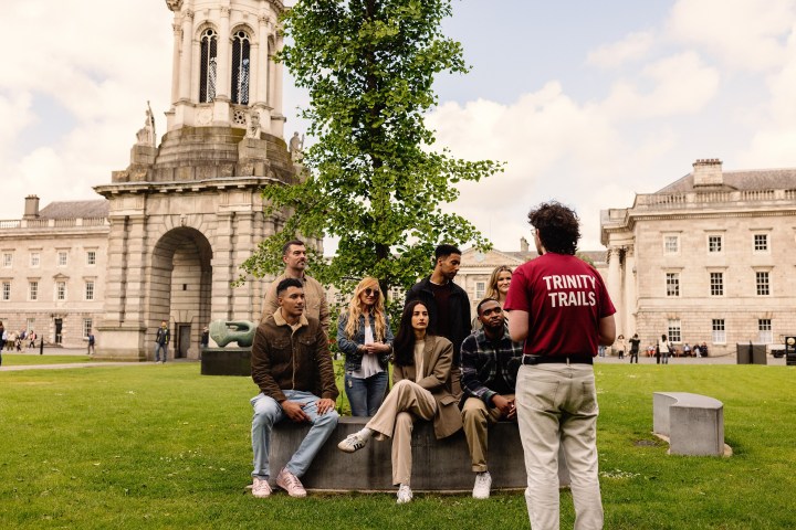 A tour guide speaks to a group who are sitting on a bench in the grass in Trinity's front square