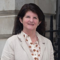 A woman with brown shoulder length hair and a beige blazer with an old stone wall behind her.