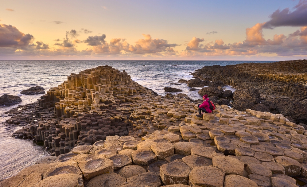 A woman in a red jacket sits on the Giants Causeway looking out to sea